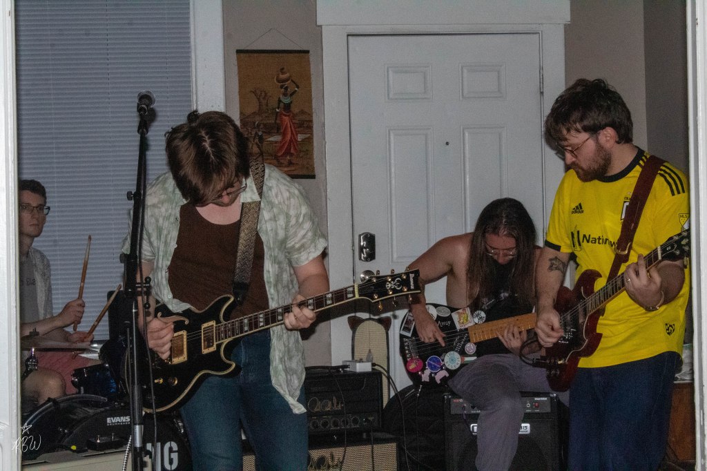 Passing On The Right play an EP release show in a living room. A man in the back left sits at a drum set, another on the right sits an amp and plays bass. In front of the bassist, stands a man playing guitar and other beside him on the left plays guitar and sings.