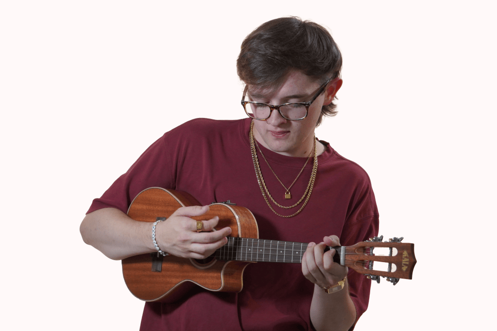 A man wearing a pair of glasses and a red shirt strums a ukulele.