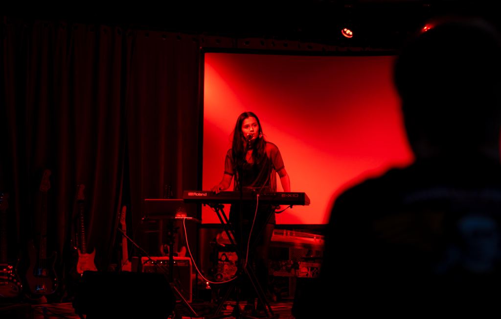A women stands in front of a keyboard and microphone in a dark room only lit by a red light.